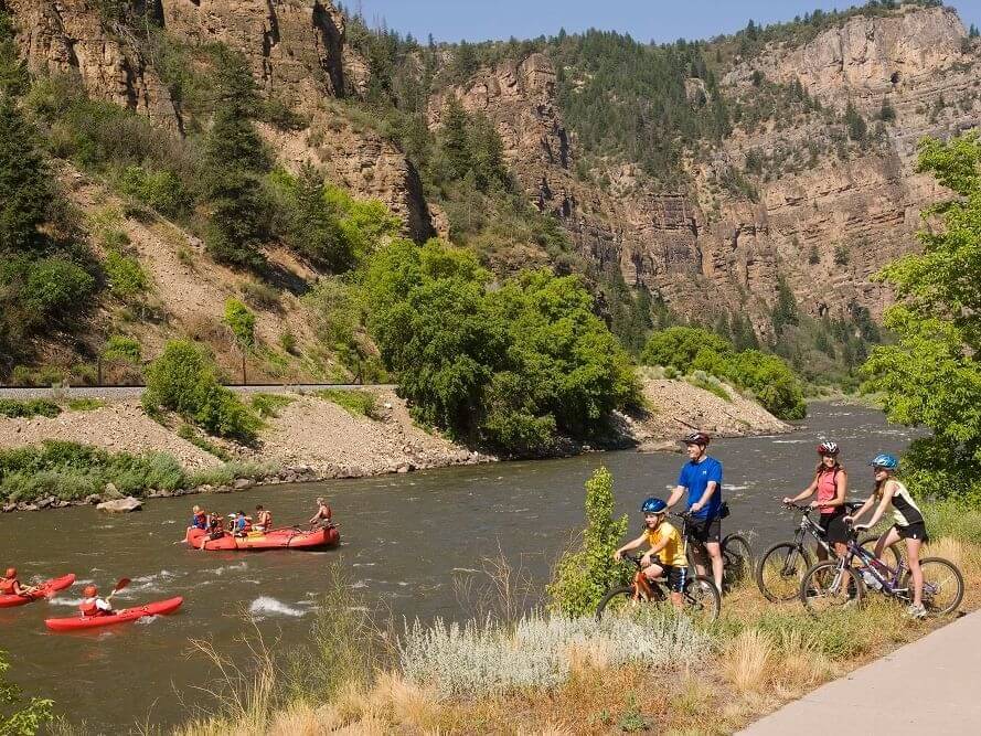 A group of cyclists pauses on a path next to the river, where people are enjoying activities like rafting. The scene unfolds against steep, rocky cliffs adorned with green trees, under a clear and sunny sky.