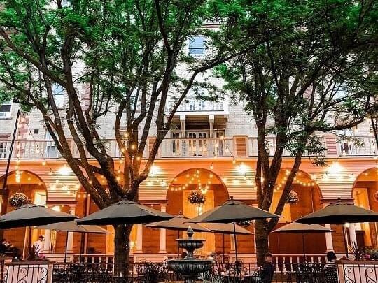 Outdoor café with large trees and string lights creating a cozy atmosphere. Melodic music drifts through the air as umbrellas cover tables near a central fountain. The building in the background has arches and hanging flower baskets, all illuminated by warm lights.