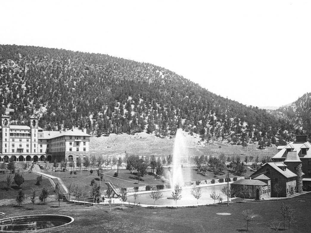 A historical black and white photo captures the stately Walter Building beside a landscaped garden featuring a tall fountain at its center, all set against a backdrop of tree-covered hills.