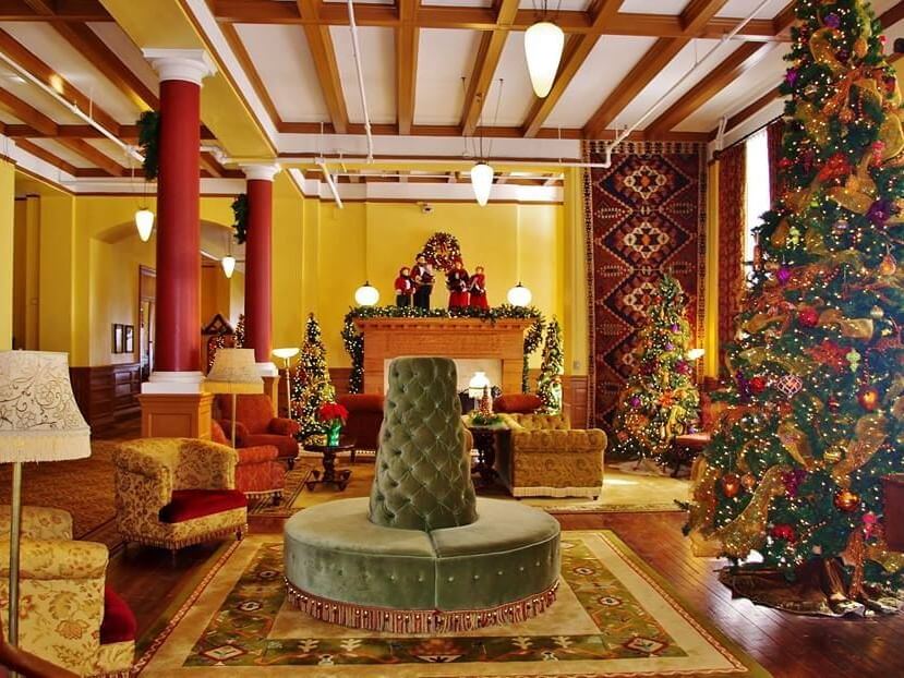A festive room celebrates December with a large Christmas tree adorned in ornaments and lights, a cozy fireplace with stockings, and plush seating, including a green circular sofa. Warm lighting enhances the holiday ambiance.