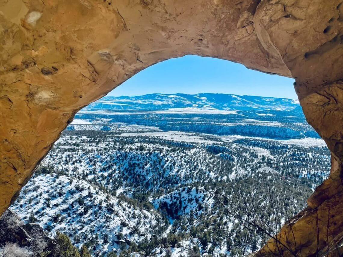 View through a rocky archway revealing an off-the-beaten-path landscape of snow-dusted hills and valleys under a bright blue sky, with distant mountain ranges on the horizon.