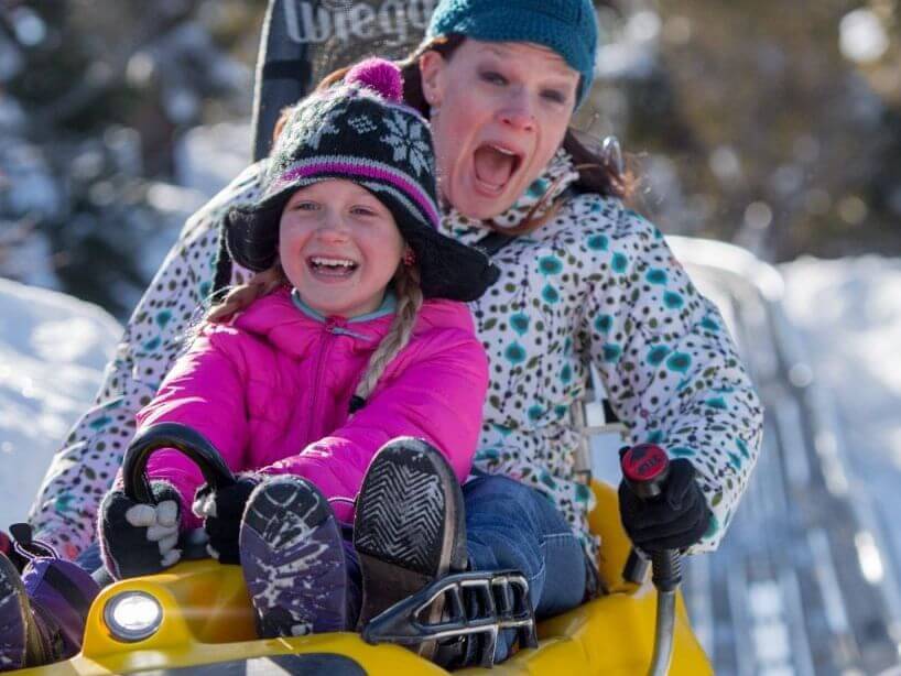 A woman and young girl, bundled in winter clothes, joyfully ride a yellow sled down a snowy slope. The girl, in her pink jacket, steers as the woman beside her looks excited. Snowy trees frame this idyllic scene, capturing the warmth and joy of being home for the holidays.