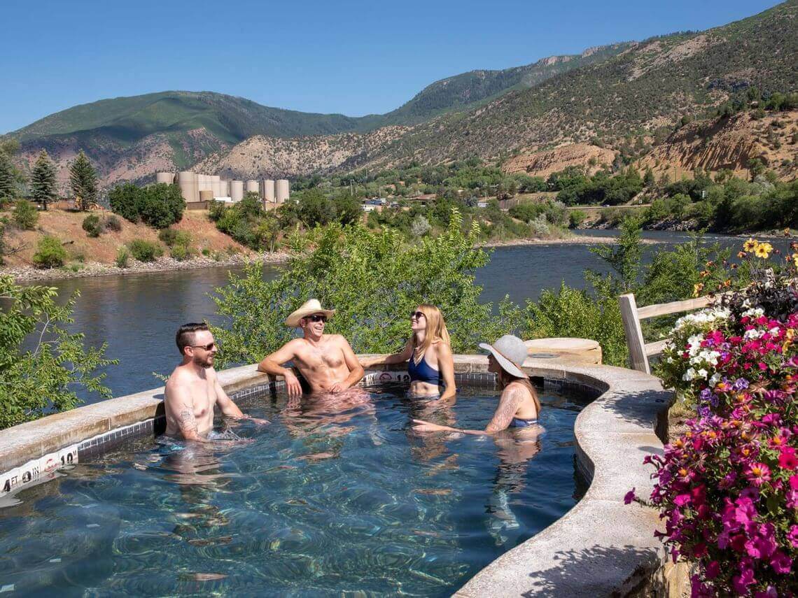 Four people relax in a hot tub overlooking a scenic river and green hills. Two wear hats, and all are in swimwear, savoring the summer sun. Bright flowers and lush trees border the hot tub area, with distant mountains framing the idyllic scene.