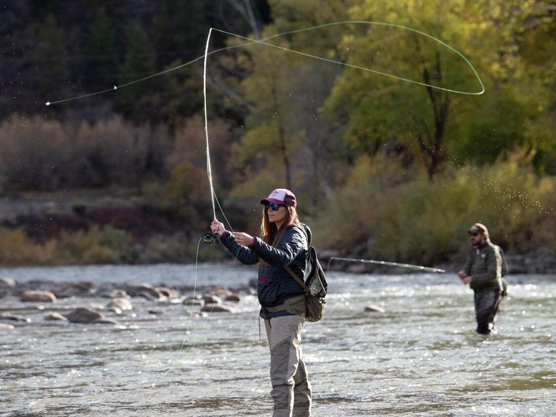 Two people fly-fishing in a river surrounded by trees with fall foliage. The person in the foreground casts a line, creating gentle ripples, while standing in the water, wearing a cap and sunglasses. In the background, another person stands further down the river amidst nature's vibrant hues.