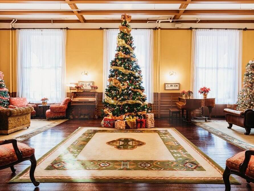 A warmly lit room features a large, decorated Christmas tree in the center, embodying the spirit of the holiday season. Vintage furniture, including armchairs and a wooden piano, surrounds it. Wrapped gifts are nestled under the tree, and sheer white curtains adorn the large windows.