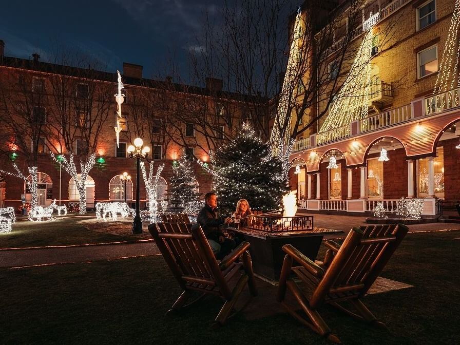 A couple sits by a fire pit in a festive courtyard adorned with strings of white lights, embracing the holiday spirit. Trees and a historic brick building are illuminated against the night sky, creating a warm and cozy atmosphere perfect for this special season.