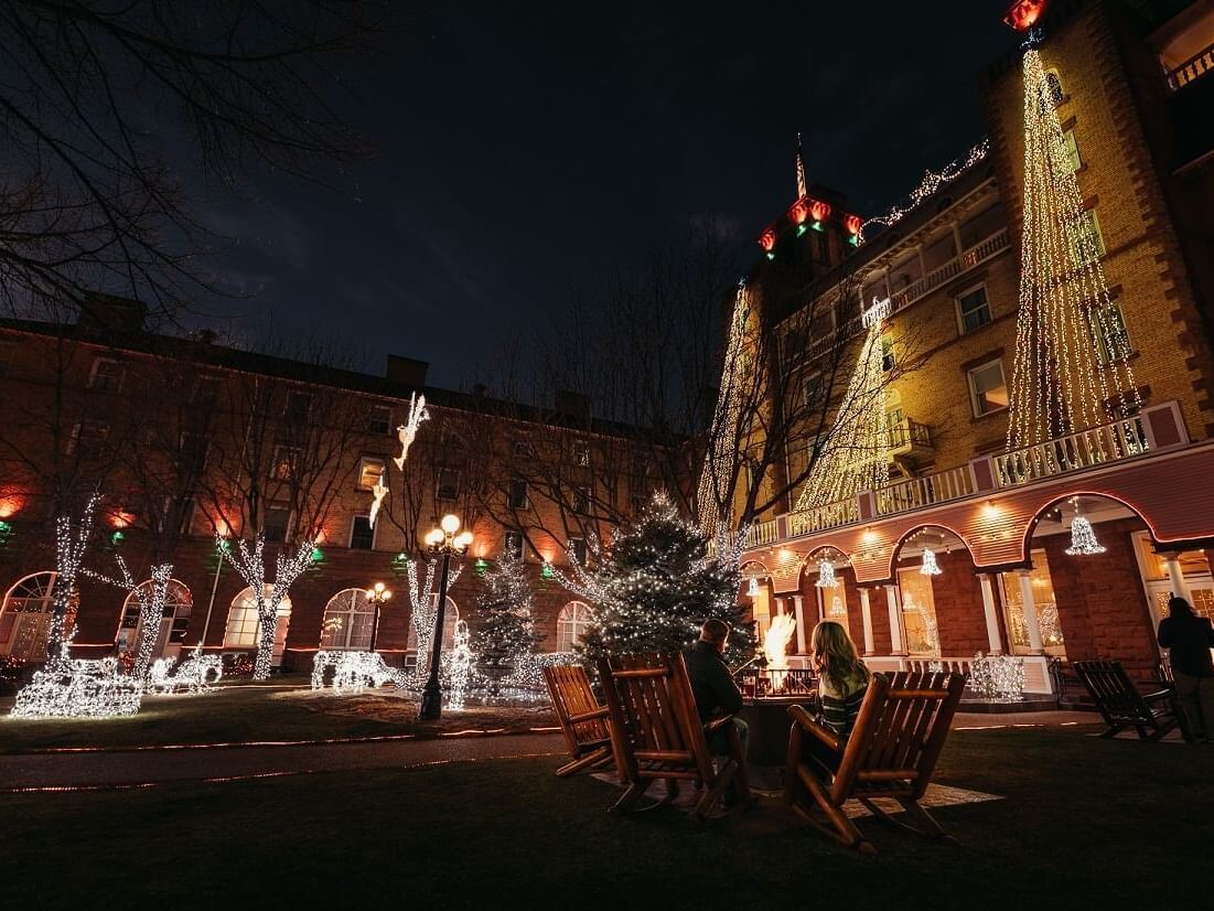 In an outdoor nighttime scene, a lit courtyard glows warmly as trees and a building are adorned with festive string lights. In the foreground, two people sit on wooden chairs near a decorated tree, creating an inviting atmosphere.