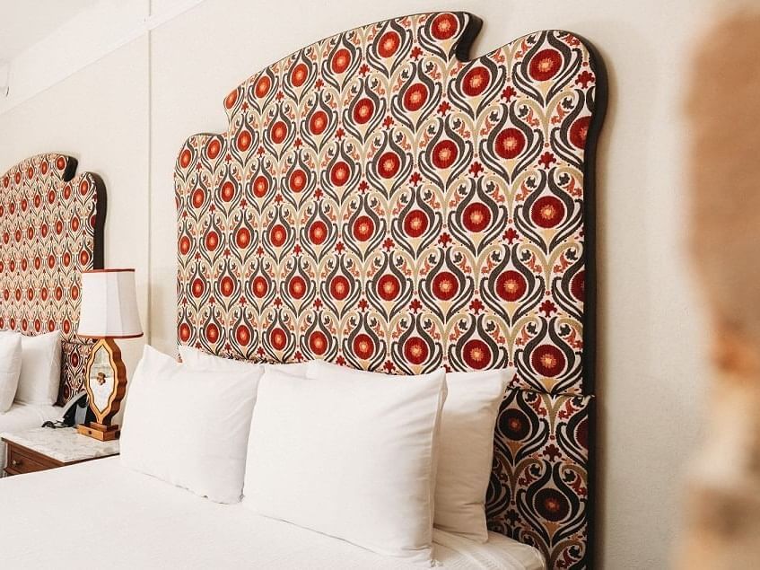 A bed with a decorative headboard featuring a colorful, intricate pattern of red, orange, and beige shapes reclaims its charm. Four white pillows are neatly arranged against it, and a nightstand with a lamp is visible beside the bed.