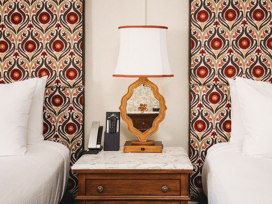 A newly renovated classic bedroom setup features two beds adorned by an ornate table lamp resting on a marble-top wooden nightstand. Two patterned curtains with red and black designs frame the lamp, while an alarm clock and phone complete the elegant scene.