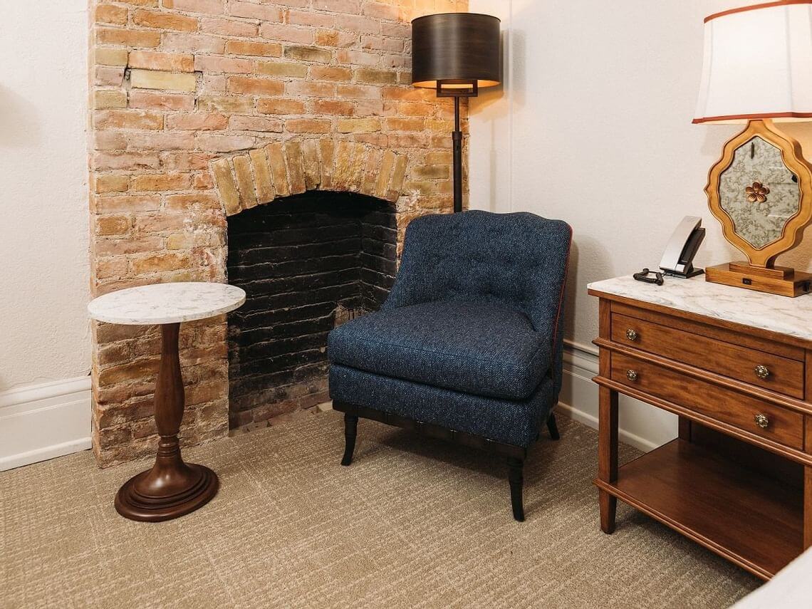 A cozy room perfect for a romantic getaway features a plush blue chair in front of a brick fireplace. A small round wooden table with a marble top sits to the left, while to the right, a wooden dresser with a lamp adds warmth. Soft carpet completes this intimate retreat.