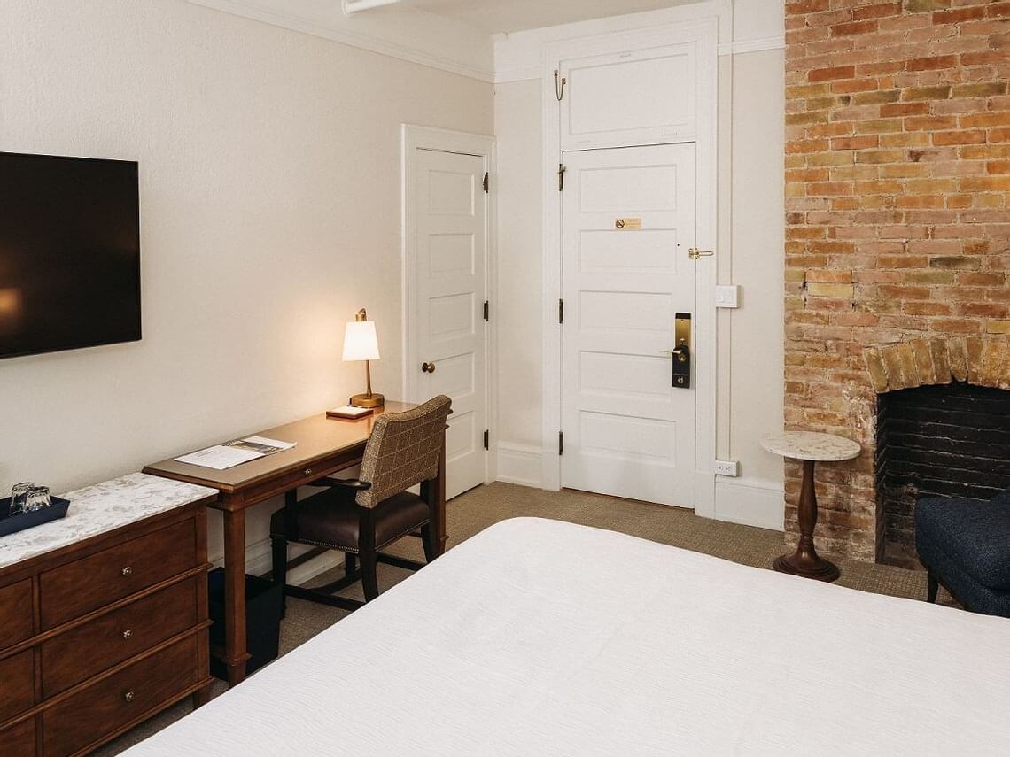 A cozy hotel room perfect for a romantic getaway, featuring a bed, a wooden desk with a lamp and chair, a mounted TV, and a brick fireplace. The room has neutral walls, white doors, and a small round side table next to an upholstered chair.