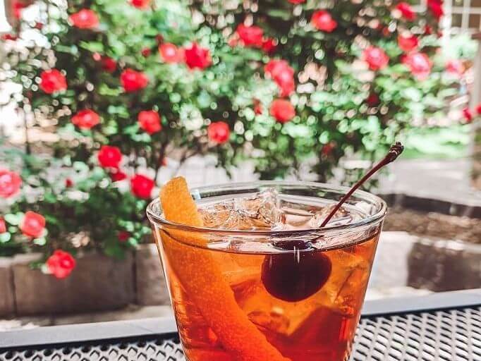 A cocktail with ice, an orange twist, and a cherry sits on a table in a garden, reminiscent of post-polo relaxation. Red flowers and greenery blur together in the background, creating a bright and refreshing outdoor scene.
