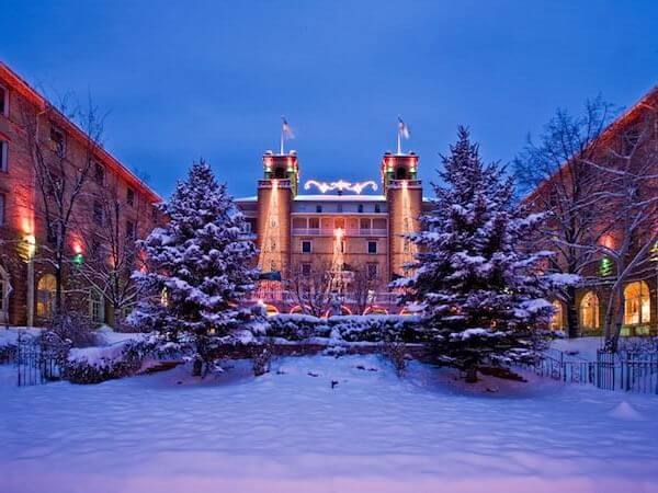 A reimagined grand building with illuminated towers stands majestically in a snowy courtyard. The scene is framed by snow-covered trees, as colorful lights glow warmly from the surrounding structures, set against a twilight sky.