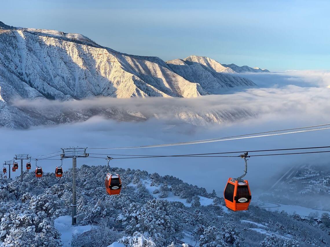 Orange cable cars transport passengers over a snow-covered mountainous landscape, offering an exhilarating outdoor adventure. Below, a layer of fog blankets the forested valley. Sunlight highlights the rugged peaks in the background, creating a picturesque winter scene.