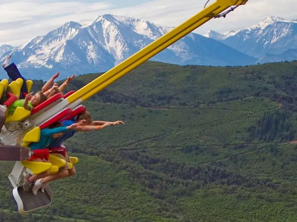 People on an adventure ride extend over a lush green valley, with snow-capped mountains in the background. They are seated in colorful harnesses with arms outstretched, savoring the view and the thrill of excitement.