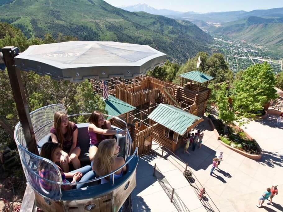 A group of people enjoy a circular amusement park ride during spring break, overlooking a scenic landscape. Below, a wooden fort-like structure and winding path are nestled among lush green hills with a distant view of the valley.