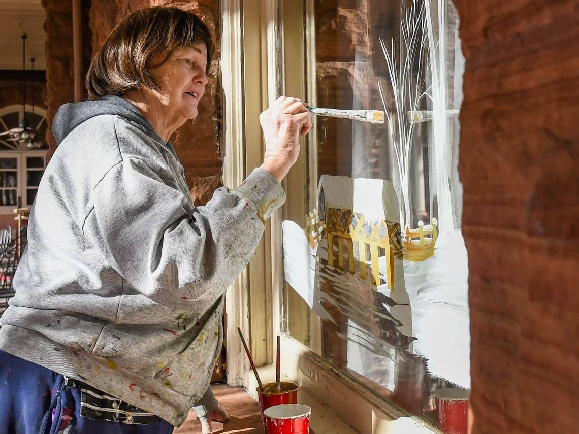 Angela Drake, in a gray hoodie, paints a winter scene on a glass window. With a brush in hand against the snowy backdrop, various paint supplies lie nearby. Sunlight bathes her as she works, capturing the essence of the moment.