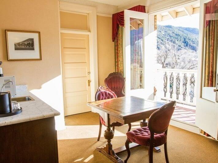 A sunlit room perfect for planning, featuring a wooden desk with two upholstered chairs. French doors open to a balcony with mountain views. A kitchenette with a coffee maker is on the left, and a framed picture hangs on the beige wall.