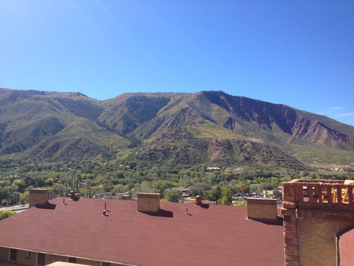 A scenic view of a range of green mountains under a clear blue sky awaits, offering a taste of the suite life. In the foreground, the roof of a building with several chimneys overlooks a lush, green valley nestled below the majestic peaks.