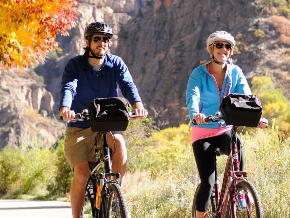 Two people, immersed in a fall color trip, are biking on a scenic path with helmets and sunglasses. Surrounded by autumn foliage and a rocky landscape, they have bags attached to their bicycles and appear to be enjoying the ride.
