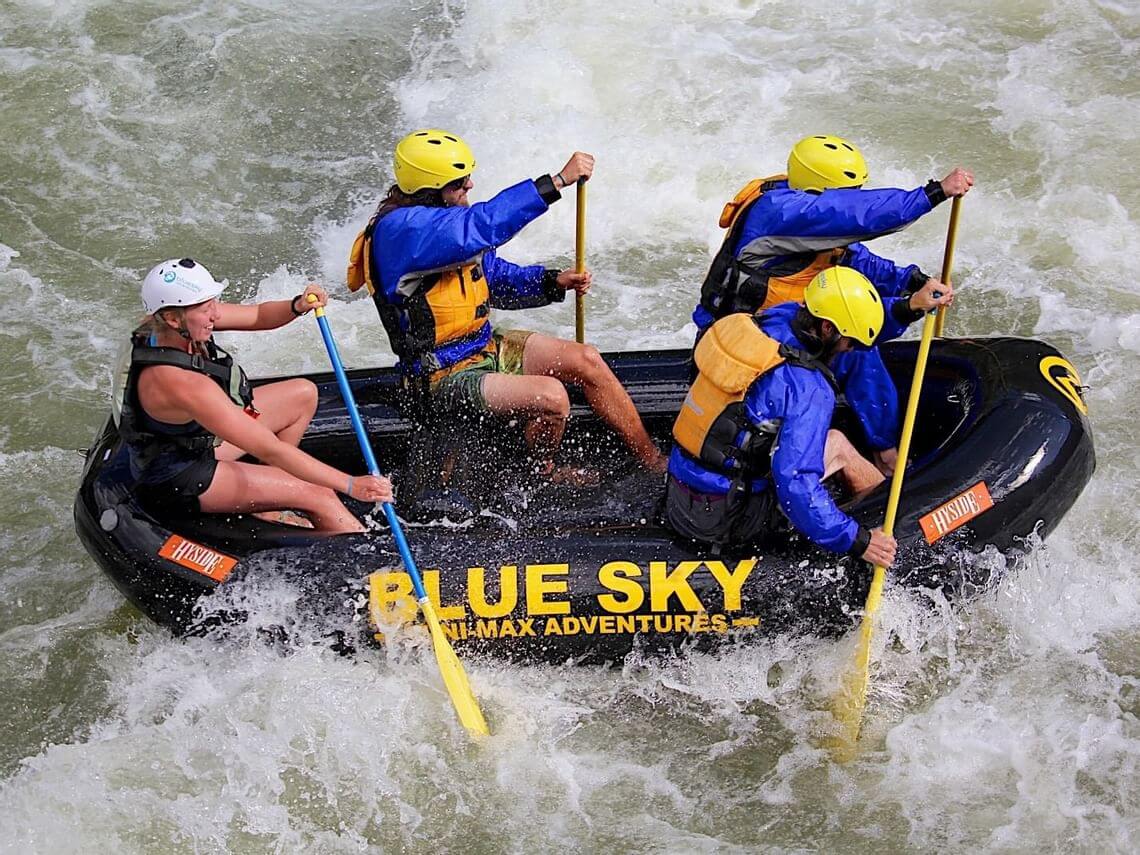 Four people in helmets and life jackets are navigating a black raft through Glenwood Canyon's river rapids. The text Blue Sky Whitewater Adventures is visible on the raft. Water splashes around as they paddle energetically.