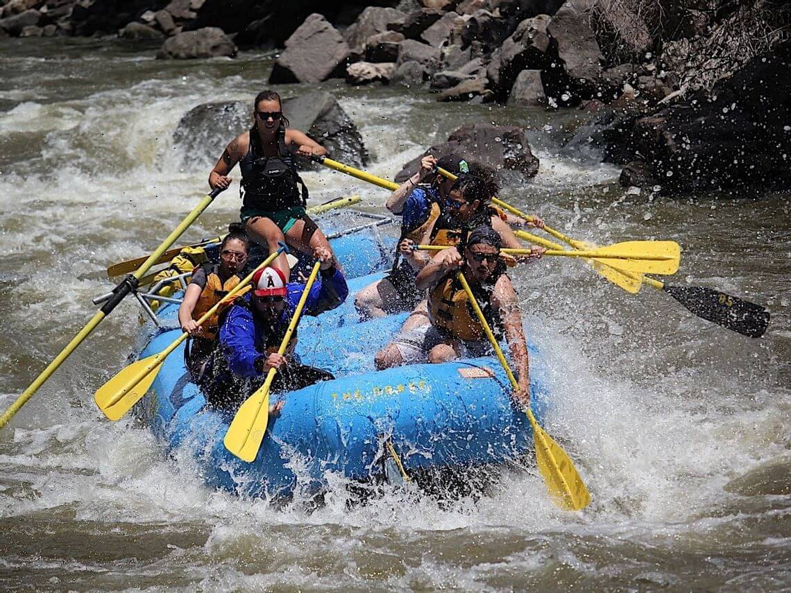 A group of six people is white water rafting on a turbulent river, experiencing one of nature’s wonders. They energetically paddle their blue inflatable raft through splashing water, set against a backdrop of rocky riverbanks—a truly dynamic and adventurous scene.