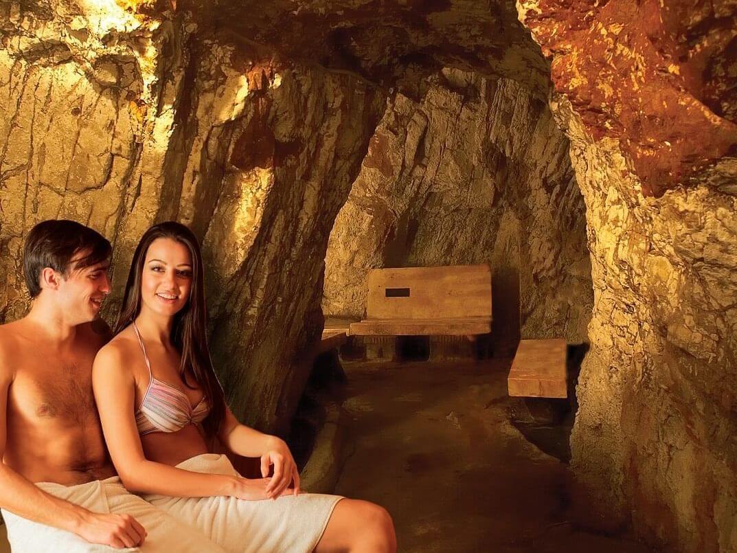 A man and woman in swimsuits sit wrapped in towels inside a rustic, warmly lit cave sauna, truly one of nature's wonders. The rocky walls and ceiling create a cozy atmosphere reminiscent of natural wonders. Benches are visible in the background.
