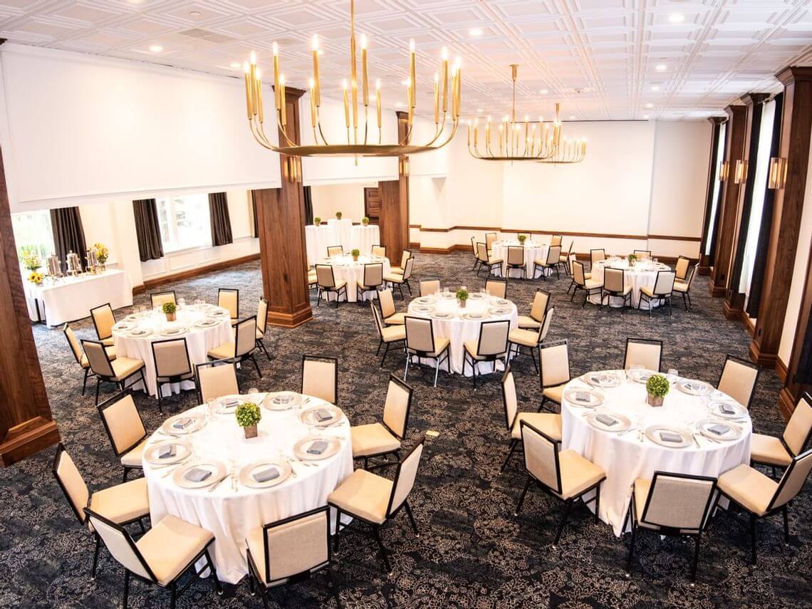 A spacious banquet hall, versatile as a meeting space, features round tables ready for an event. Each table is draped with a white tablecloth and adorned with a small plant centerpiece. Elegant chandeliers hang from the ceiling, and large windows are framed with curtains.