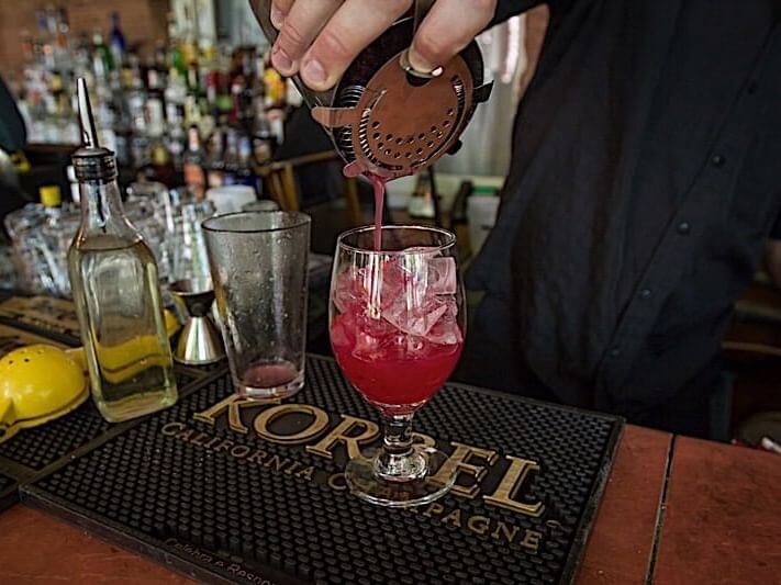 A bartender pours a red cocktail into a glass with ice using a strainer, as sunlight filters through the courtyard. The scene includes a bar counter adorned with various drink-making tools, bottles, and a black bar mat labeled Korbel California Champagne.