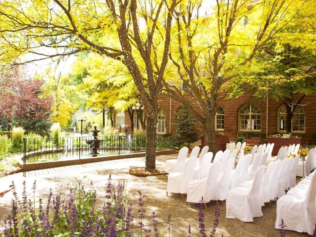 An enchanting outdoor fall wedding setup features white chairs arranged in rows beneath vibrant yellow-leaved trees. A brick building with arched windows stands proudly in the background, while a small fountain nestles among the lush greenery.