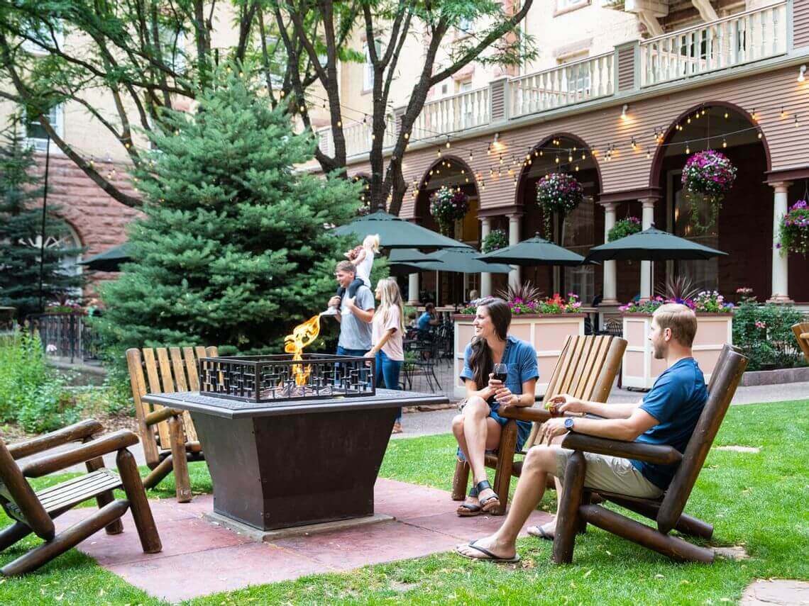 People relax in wooden chairs around a fire pit in a lush courtyard with a historic building, basking in the summer warmth. A couple enjoys drinks, while others, including a child, walk nearby. Hanging flower baskets and trees enhance the tranquil setting.