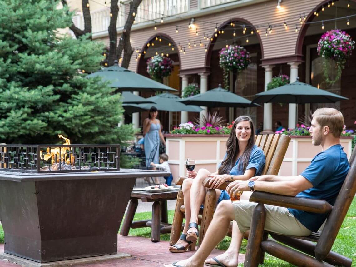 A couple sits on wooden chairs around a fire pit in a lush garden, enjoying drinks during their romantic retreat. String lights and large arched windows adorn the background, with hanging flower pots adding to the ambiance. An umbrella-covered table and a woman standing nearby complete the scene.