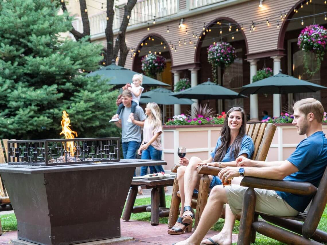 A group of people relax outdoors by a fire pit at a cafe. A couple sits on wooden chairs, enjoying drinks, while romance sparks in the air. Nearby, a man carries a child through the lush greenery. Flower pots and string lights enhance the cozy atmosphere.