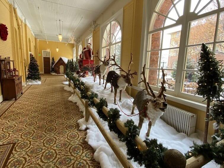 A festive hallway display reimagines Santa's sleigh and reindeer surrounded by artificial snow. Decorated trees line the path, while large windows let natural light fill the space, creating a magical winter wonderland.