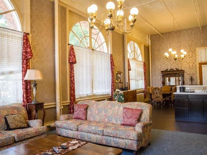 Experience the suite life in this spacious, ornate living room adorned with high ceilings and elegant chandeliers. The room boasts large arched windows, floral-patterned sofas with cushions, a wooden coffee table, and a dining area highlighted by a vintage cabinet in the background.