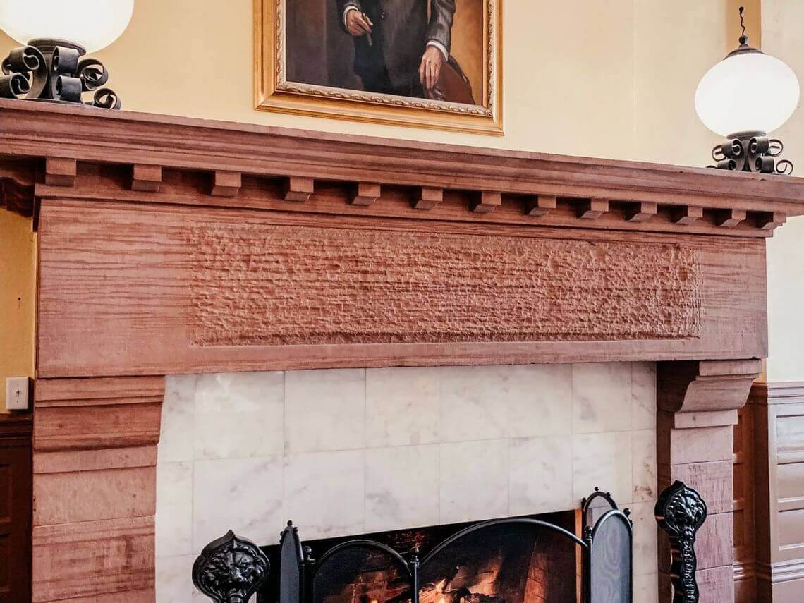 A vintage fireplace with a carved wooden mantelpiece, flanked by ornate metal tools. Above hangs a framed portrait of Walter. Two spherical lamps rest on the mantel, illuminating the scene.