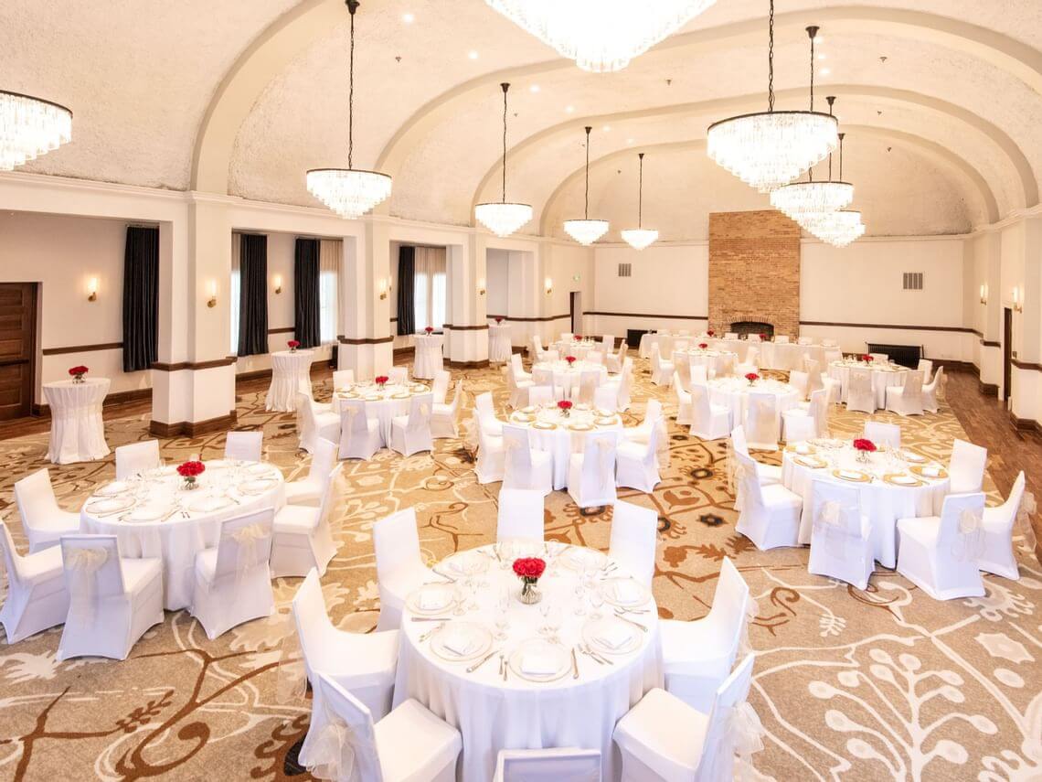 Elegant meeting space with round tables covered in white tablecloths, set with dishes and red floral centerpieces. The room features high ceilings, chandeliers, a large carpet with floral patterns, and a stone fireplace at one end.