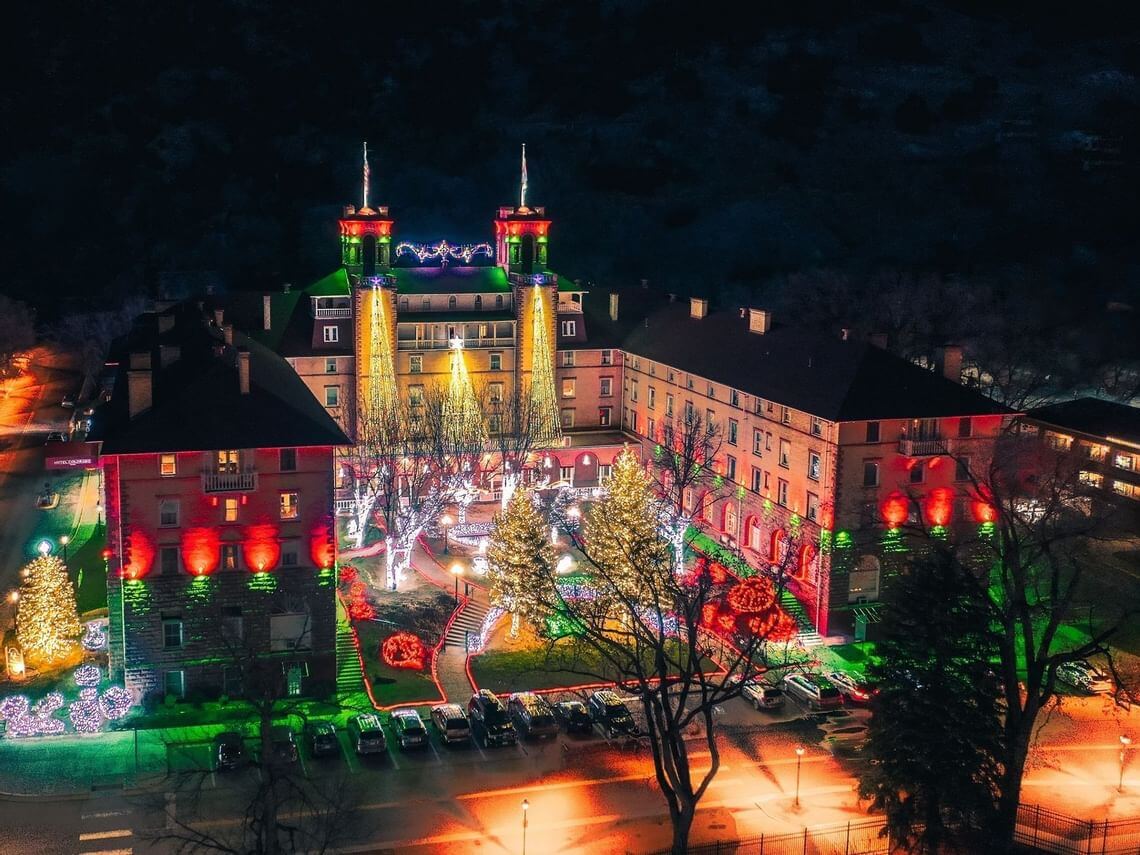 A large building is beautifully illuminated with colorful lights in a festive holiday display at night. Trees and shrubs are decorated with white and multi-colored lights, while parked cars line the street in front.