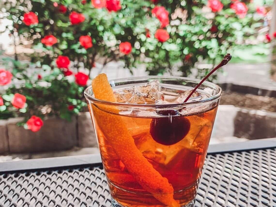 A glass of cocktail with an orange peel and cherry garnish sits on a metal table, perfectly complementing the vibrant courtyard where red flowers are in full bloom.