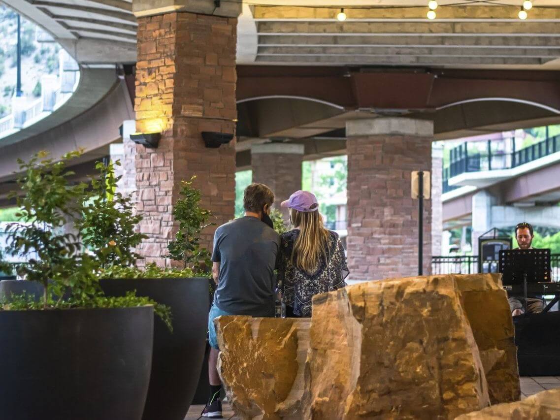A couple sits on a bench in an outdoor area under a bridge, surrounded by large planters and stone decorations. The ripple of the musician's melody in the background creates a tranquil atmosphere, while hanging lights add a warm glow to the scene.