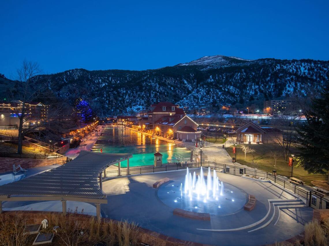A scenic view of a resort at dusk features a large pool inviting guests to soak away their worries. The pool is flanked by buildings with illuminated windows, while a fountain graces the foreground. Snow-capped mountains and softly lit trees enhance the tranquil atmosphere.