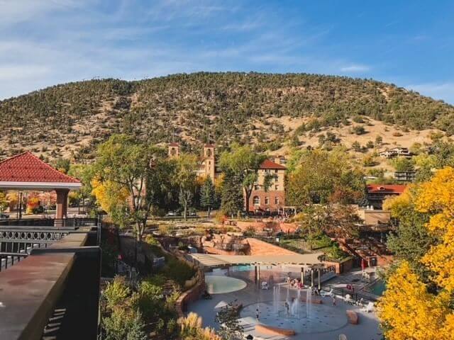 A scenic view of a park with a fountain, surrounded by vibrant fall trees. In the background, a lush hill rises under a clear blue sky. Buildings with red roofs are nestled among the autumn foliage.