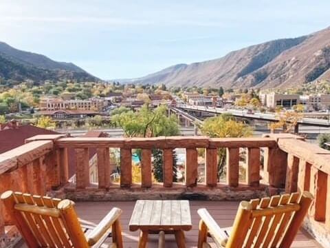 A scenic view from a wooden balcony with two chairs and a small table overlooks a town nestled between mountains. Vibrant fall colors paint the trees, as buildings stretch across the valley under a clear blue sky.