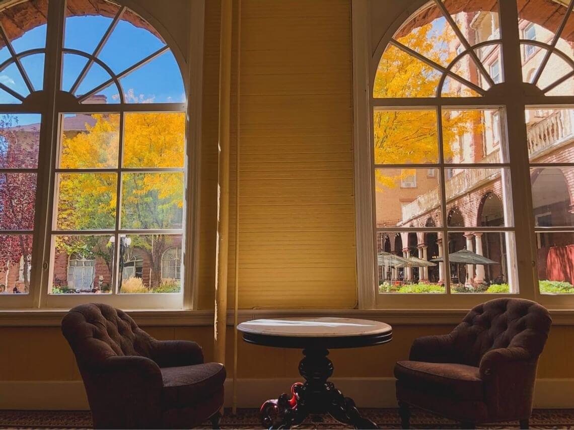 Two plush chairs and a round table by large windows frame a vibrant fall scene outside. Yellow leaves on trees contrast with the reddish-brown brick building, while the bright blue sky enhances the warm, cozy interior ambiance.