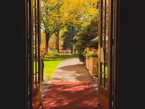 View of a picturesque garden path through open double doors, framed by vibrant fall colors and lush greenery. A red carpet stretches outside, with sunlight casting warm tones over the scene.