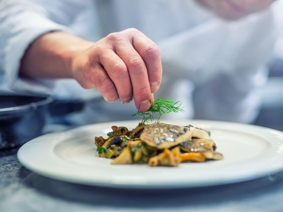 A chef garnishes a plated dish with fresh dill. The fresh dish, featuring mushrooms and a fillet, is artfully arranged on a white plate, while the chef's hand is in focus, emphasizing the finishing touch.