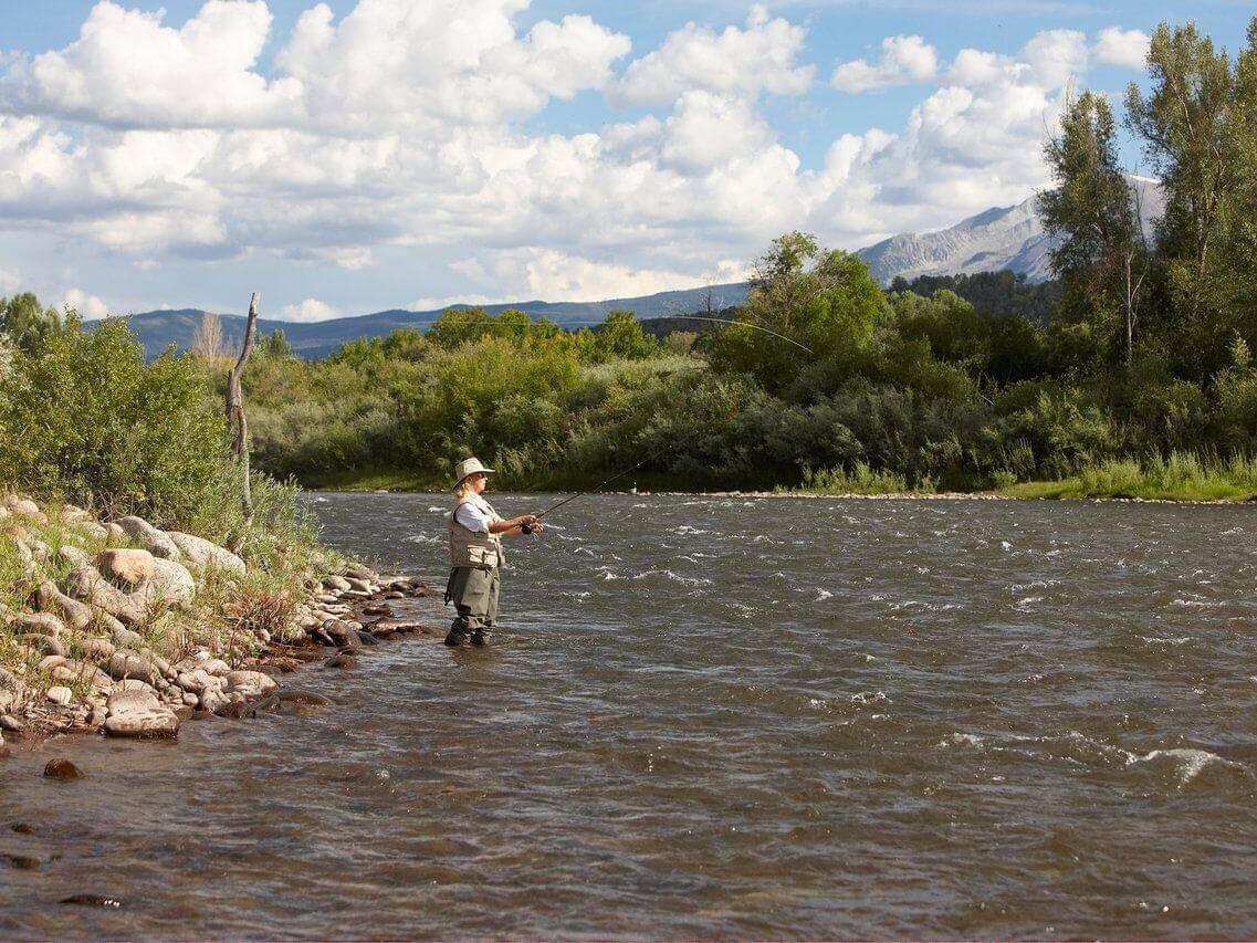 A person fly fishing in a river stands near the rocky shoreline, surrounded by lush green trees and distant mountains under a sky filled with fluffy white clouds—a perfect summer day.