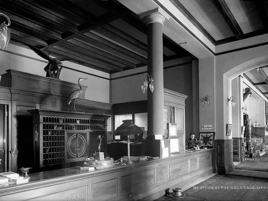 A black and white image of a vintage hotel lobby reveals a wooden front desk with decorative columns, linked to its era. Taxidermy birds perch above while papers are scattered on the desk. An archway gracefully leads to another room in the background.