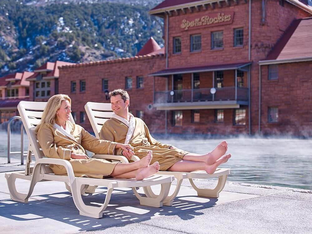 A couple in robes blissfully relaxes on lounge chairs beside a steaming outdoor pool. In the background, a large brick building labeled Spa of the Rockies stands majestically against a mountainous landscape.
