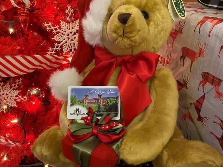 A teddy bear with a red bow and Santa hat sits holding a gift box with a bow. In front is a gift card displaying a building and the words A Gift for You. The background features a red Christmas tree adorned with candy cane decorations, perfect for enjoying the best coffee this festive season.